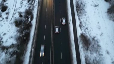 Karla kaplı bir yol boyunca giden arabaların hava görüntüsü, aracın hareketini ve kışın etrafındaki manzarayı gösteriyor, kamera arabaları sorunsuzca takip ediyor. Otomobiller soğuk mevsimde otoyolda gidiyor.