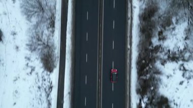 Karla kaplı bir yol boyunca giden kırmızı bir arabanın hava görüntüsü aracın hareketini ve etrafındaki kış manzarasını gösteriyor. Kamera arabayı sorunsuz bir şekilde takip ediyor.