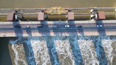 Köprüde akan su ve araçların yer aldığı nehir üzerindeki barajın hava perspektifi, kademeli olarak yakınlaştırarak sürekli eylem ve sahne gelişimini vurguluyor.