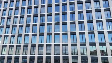 Modern ofis binasının dış görünüşü gökyüzünü ve çevreyi yansıtan bir dizi büyük pencereyi gösteriyor, mimari tasarım ve yapısal unsurları sürekli bir bakış açısıyla vurguluyor.