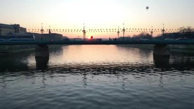 Gün batımında nehrin üzerindeki köprüden geçen tramvayın hava görüntüsü arabaların yanından geçişi, dinamik hareketler ve akşam gökyüzünün canlı renkleri suya yansıyor.