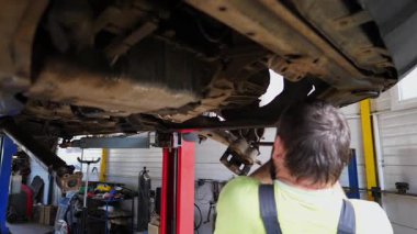 Üniformalı otomobil tamircisi, iş yerinde ingiliz anahtarı kullanarak arabanın tamirini yapıyor. Profesyonel erkek tamirci garajdaki bir kaldırma aracının altında çalışıyor. Otomobil bakımı kavramı.