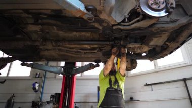 Üniformalı araba tamircisi atölyedeki ingiliz anahtarı aletini kullanarak arabanın tamirini yapıyor. Profesyonel erkek tamirci garajdaki bir kaldırma aracının altında çalışıyor. Otomobil bakımı kavramı.