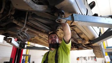 Yetişkin profesyonel tamirci serviste ya da garajda araba tamir ediyor. Erkek oto tamircisi atölyede kaldırma aracının altında çalışıyor. Düzenli otomobil bakımı kavramı. Yavaşça kapat..