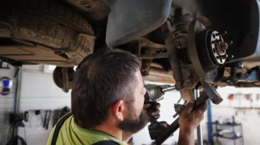 Üniformalı profesyonel sakallı tamirci atölyede çekiç kullanan arabanın tamirini yapıyor. Yetişkin erkek tamirci garajdaki bir kaldırma aracının altında çalışıyor. Otomobil bakımı kavramı.