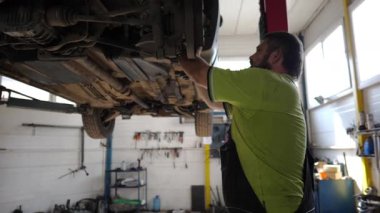 Üniformalı araba tamircisi atölyede ingiliz anahtarı kullanarak araba tamiri yapıyor. Yetişkin erkek tamirci garajdaki bir kaldırma aracının altında çalışıyor. Otomobil bakımı kavramı.