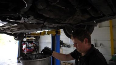 Profesyonel tamirci üniformalı. Garajdaki arabanın altındaki kirli yağı boşaltıyor. Genç tamirci atölyede kaldırma aracının altında çalışıyor. Otomobil bakım kavramı.