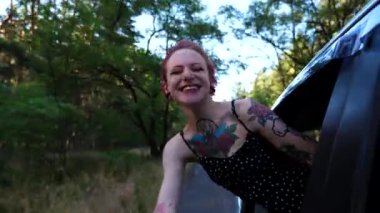 Araba camından sarkan dövmeli mutlu punk kızı ve kır yolunda gezintiden zevk alıyor. Pembe saçlı neşeli hippi kadın araba yolculuğunda eğleniyor. Seyahat konsepti. Yavaş çekim.