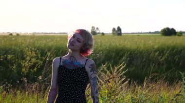 Kırsal kesimde fotoğraflara poz veren dövmeli kaygısız serseri kız. Pembe saçlı mutlu bayan hipster gün batımında kırsal kesimde fotoğraflanır. Hippi kadın doğada dinleniyor. Kapat..