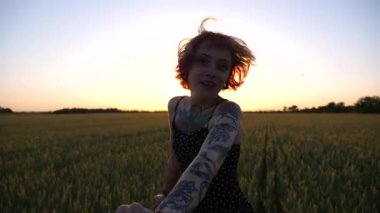 Gün batımında yeşil buğday tarlasında koşan erkek elini tutan mutlu punk kızı çek peşimden. Neşeli bayan hippi, erkek arkadaşını arpa çayırlarının arasına çekiyor. Mutlu çift doğayla eğleniyor. POV.