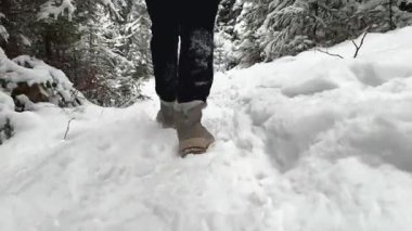 Genç bir kadın kış botlarıyla karlı orman yolunda yürüyor, taze kardaki ayak seslerini yakalıyor, uzun yeşil ağaçlar ve sakin bir kış manzarasıyla çevrili.