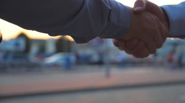 Gün batımında iki kendine güvenen meslektaşın şehir sokağında el sıkışırken çekilmiş. Başarılı genç iş adamları, araba parkının arka planında birbirlerini selamlıyorlar. Yavaşça kapat..