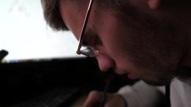 Erkek komisyoncu, bilgisayar ekranında döviz grafiklerini analiz ediyor ve kağıda veri yazıyor. Finans analisti akşam vakti borsada işlem yapıyor. Bitcoin BTC kavramı. Yavaş çekim.