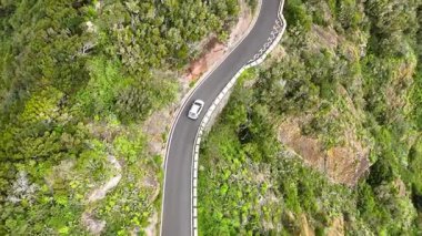 Bereketli yeşil tepeler ve kayalık uçurumlarda dolambaçlı bir yolda giden beyaz bir arabanın hava görüntüsü. Otomobiller manzarada geziniyor. Otomobiller güzel bir rotada gidiyor. Yaz gezisi