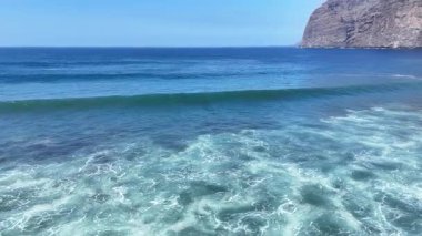 Okyanus dalgalarının kıyıya vuruşunu gösteren hava manzarası, berrak mavi gökyüzünün altındaki kayalık bir uçuruma karşı suyun ve köpüğün dinamik hareketini gösteriyor. Yuvarlanan deniz dalgaları