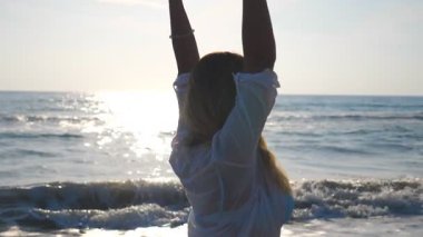 Bikinili ve tişörtlü genç bir kadın kıyıda dönüyor ve ellerini kaldırıyor. Sarı saçlı güzel kız deniz kıyısında yürüyor ve hayatın tadını çıkarıyor. Yaz tatilinde rahatla. Yavaşça kapat..