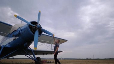 Pilot mavi çift kanatlı uçakla etkileşime girer, pervane ve motoru inceler, titiz uçuş öncesi kontrolleri ve havacılık güvenliği ve performansına bağlılığı vurgular.