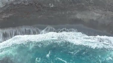 Okyanus dalgaları koyu kumlu sahile çarpar, köpük ve dalgalanmalar yaratır, önplanda berrak mavi su gradyanı ve arka planda kayalık kıyı şeridi görülür. Siyah volkanik kum. Hava görünümü