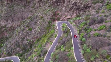 Kırmızı araba keskin virajlar ve yemyeşil yemyeşil ile dolambaçlı dağ yolunda yol alır. Kayalık uçurumlar ve canlı bitki örtüsü olan manzaralı bir yolda araba sürmek. Güzel doğa rotasında yolculuk. Hava görünümü