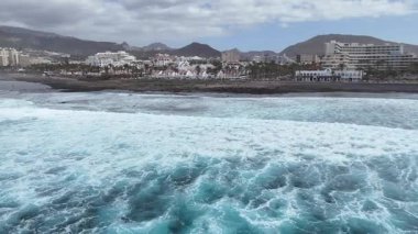 Canlı bir sahil sahili sahili sahili. Dalgalar kayalara çarpıyor. Palmiye ağaçları, oteller ve dağlar tropikal bir ortamda parçalı bulutlu bir gökyüzü altında sergileniyor. Hava görünümü