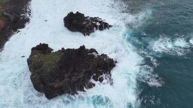 Kaya kıyı şeridinin çarpışan dalgalarla hava görüntüsü, okyanus suyu ve engebeli kıyı arasındaki dinamik etkileşimi gösteriyor, kıyı manzarasının doğal güzelliğini vurguluyor.