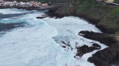 Dalgalar kıyıya yuvarlanan köpüklü sörflerle kayalık sahil şeridine çarpar ve bulutlu gökyüzünün altındaki arka plandaki kayalıklara tünemiş renkli binalar ortaya çıkar. Hava görünümü