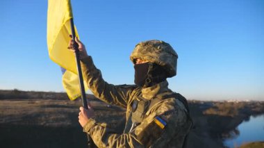 Askeri üniformalı erkek askerin Dolly çekimi Ukrayna bayrağını sallayarak tepenin zirvesinde duruyor. Ukraynalı ordu mensubu, günbatımının arka planında ulusal bayrağı kaldırdı. İstila direnişi kavramı.