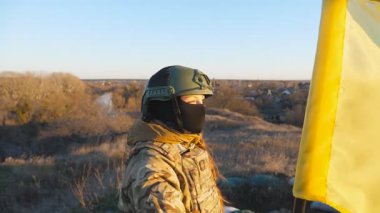 Dolly 'nin kadın asker çekimi Ukrayna bayrağını sallayarak tepenin zirvesinde duruyor. Kamuflaj üniformalı genç Ukraynalı asker gün batımında mavi-sarı pankart kaldırıyor. İstila direnişi kavramı.