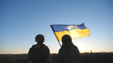 Ukrayna ordusunun kadın ve erkek askeri tepenin zirvesinde mavi-sarı bayrak sallıyor. Askeri üniformalı genç kadın ve adam gün batımına karşı Ukrayna bayrağı kaldırdı. Savaşta zafer. Arka görünüm.