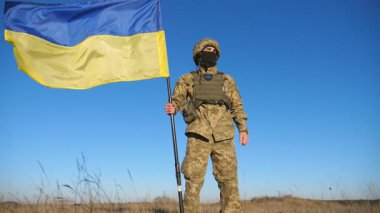 Askeri üniformalı Ukraynalı asker ulusal bayrağı kaldırıyor. Genç erkek asker mavi gökyüzüne karşı Ukrayna bayrağı sallıyor. Rus saldırısına karşı zafer. İstila direnişi. Düşük görünüm.