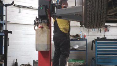 Üniformalı bir tamirci atölyede ya da garajda araba arızalarını tamir ediyor. Araba servisinde kaldırma aracıyla çalışan profesyonel bir tamirci. Düzenli otomobil bakımı kavramı. Yavaş çekim.