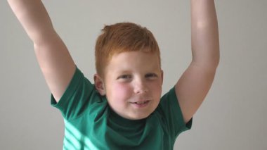Sevimli yakışıklı çocuk el kaldırıyor ve dans ediyor. Neşeli bir başarı. Mutlu genç çocuk yüzünde neşe duygularıyla kameraya bakıyor. Evde çilli bir çocuğun portresi. Yavaş çekim. Yakın çekim..