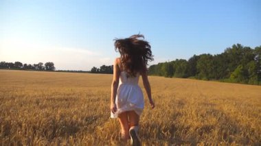 Genç kız mavi gökyüzünün altında buğday tarlası boyunca koşuyor. Kadın çayırda koşuyor. Yan görünüm Yavaş çekim.