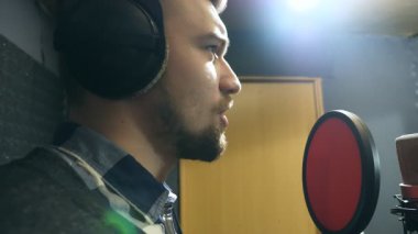 Ses stüdyosundaki mikrofona şarkı söyleyen kulaklıklı erkek şarkıcı profili. Genç adam yeni bir melodi ya da şarkı kaydediyor. Yaratıcı bir müzisyenin çalışması. Gösteri dünyası konsepti. Yavaş çekim.
