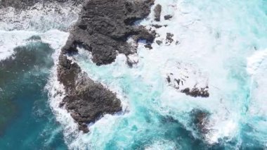 Turkuaz okyanus dalgaları kayalık kıyı şeridine çarparak, canlı mavi su ve kıyı manzarasında görünen beyaz sörf ile köpük ve sıçrama yaratırlar. Denize açılan hava manzarası. Canlı sahil manzarası