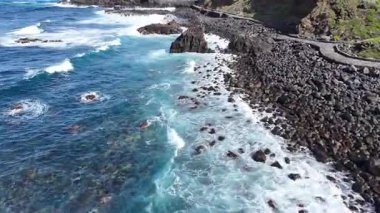 Kayalık kıyı şeridini ve çarpışan dalgaları barındıran kıyı manzarası, arka planda yemyeşil tepeler ve yukarıda açık mavi gökyüzü, doğa elementlerinin dingin güzelliğini yakalıyor. Denize açılan hava manzarası