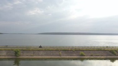 Göl kenarındaki yolda, arka planda güneş ışığı olan modern bir motosiklet süren bir adam. Motosikletli baraj yolunda motosikletiyle yarışıyor. Yolculuk sırasında bisiklet süren adam. Özgürlük kavramı. Hava görüntüsü.