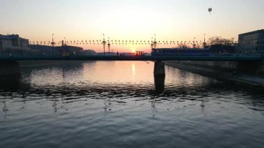 Gün batımında nehrin üzerindeki köprüden geçen tramvayın hava görüntüsü arabaların yanından geçişi, dinamik hareketler ve akşam gökyüzünün canlı renkleri suya yansıyor.