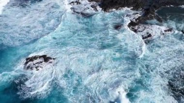 Turkuaz okyanus dalgaları kayalık kıyı şeridine çarparak köpük ve su sıçratır. Su yüzeyinin dinamik hareketinin havadan görüntüsü. Güzel renkli deniz manzarası. Kapat.