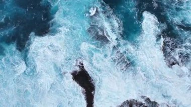 Kayalık kıyı şeridine çarpan okyanus dalgalarının hava görüntüsü, zıt kayalarla dolu canlı bir mavi deniz ortamında suyun ve köpüğün dinamik hareketini gösteriyor. Üst manzaradan deniz manzarasına