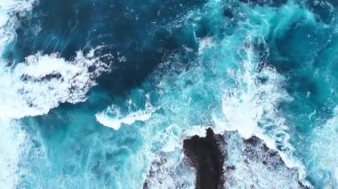 Kayalık kıyı şeridine çarpan okyanus dalgalarının hava görüntüsü, zıt kayalarla dolu canlı bir mavi deniz ortamında suyun ve köpüğün dinamik hareketini gösteriyor. Üst manzaradan deniz manzarasına