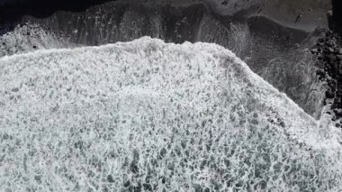 Okyanus dalgaları kayalık bir sahile çarpar, köpük ve su sıçratır, güneş banyosu yapanlar ve renkli havlular kıyı şeridinde parlak güneş ışığı altında görünür. Hava görünümü