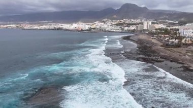 Kıyı dalgalarının dalgaların kayalık kıyılara çarptığı, sörfçülerin dalgalara bindiği ve şehrin ufuk çizgisinin bulutlu gökyüzünün altında görülebildiği hava manzarası. Turistik bir hava. Deniz kıyısının üst görüntüsü