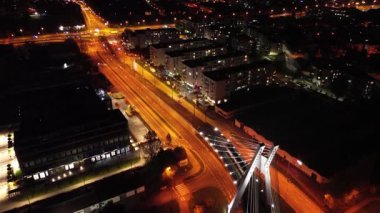 Tramvay gece modern bir ulaşım kavşağında sorunsuzca süzülüyor, şehir ışıklarıyla aydınlatılıyor, dinamik hareketler ve şehir atmosferi sergiliyor, kamera sahnenin diğer tarafına geçiyor.