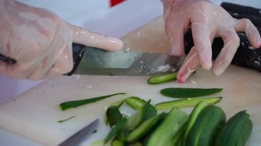 Eldivendeki yetenekli eller, taze salatalıkları kesme tahtasıyla dilimliyor. Mutfak hazırlıklarında kullanılan tekniği canlı renklerle vurguluyorlar.