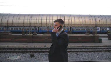 Başarılı bir iş adamı tren istasyonunda yürüyor ve telefonla konuşuyor. Demiryolu yakınlarında gezinen ve iş konuşması yapan yakışıklı bir adam. Adam dışarı çıkıp cep telefonuyla konuşuyor. Yavaş çekim.