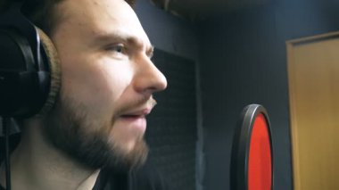 Kulaklıklı erkek şarkıcı ses stüdyosundaki mikrofona şarkı söylüyor. Genç adam duygusal olarak yeni bir melodi ya da şarkı kaydediyor. Yaratıcı bir müzisyenin çalışması. Gösteri dünyası konsepti. Yavaş çekim.