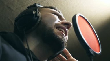 Kulaklıklı erkek şarkıcı ses stüdyosundaki mikrofona şarkı söylüyor. Genç adam duygusal olarak yeni bir şarkı kaydediyor. Yaratıcı bir müzisyenin çalışması. Gösteri dünyası konsepti. Yavaş çekim Düşük açı.