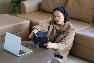 Asyalı kadın stresli bir şekilde oturup siyah bir not defterindeki bilgilere bakıyor çünkü bilgisayarıyla online çalışmakta zorlanıyor. çalışma alanı, evden çalışma