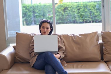 Kanepede oturan Asyalı kadın bilgisayarını internet işi için kullanıyor ya da evden çalışıyor..